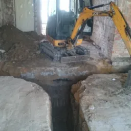 Żółta mini koparka JCB w trakcie wykonywania wykopu wewnątrz budynku z ceglanymi ścianami, widoczny usypany grunt i głęboki rów.