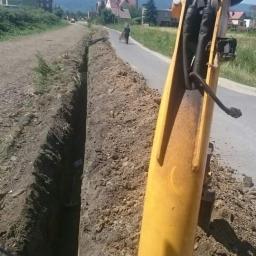 DAR-KOP - Żółta ramię koparki widoczna z bliska, wykopany wąski rów wzdłuż drogi asfaltowej, w tle zabudowania wiejskie i zalesione wzgórza, osoba z wózkiem oddalona.