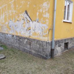 DAR-KOP - Żółta ściana budynku z kamienną podmurówką i odpadającym tynkiem, pokryta częściowo graffiti, widoczny rów odpływowy i trawa.