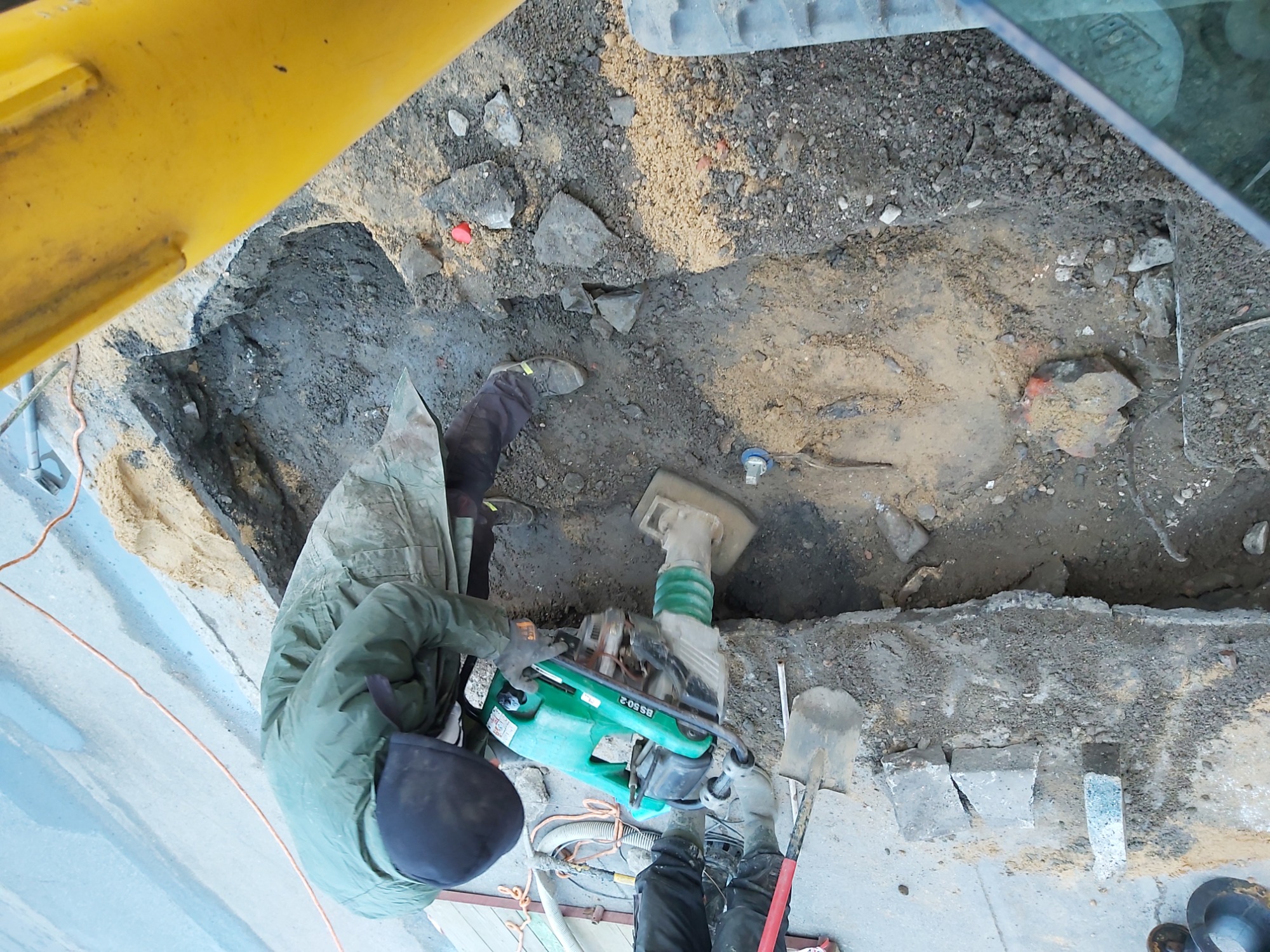 Pracownik w zielonej kurtce zagęszcza grunt w wykopie za pomocą zielono-szarej zagęszczarki skoczkowej, widok z góry z perspektywy kabiny maszyny budowlanej.