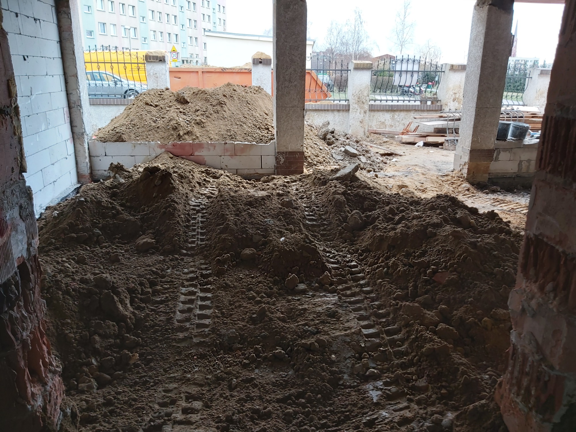 Wnętrze budynku w trakcie robót ziemnych, z hałdą ziemi i śladami opon maszyn budowlanych, widoczne elementy konstrukcyjne budynku oraz ogrodzenie w tle.