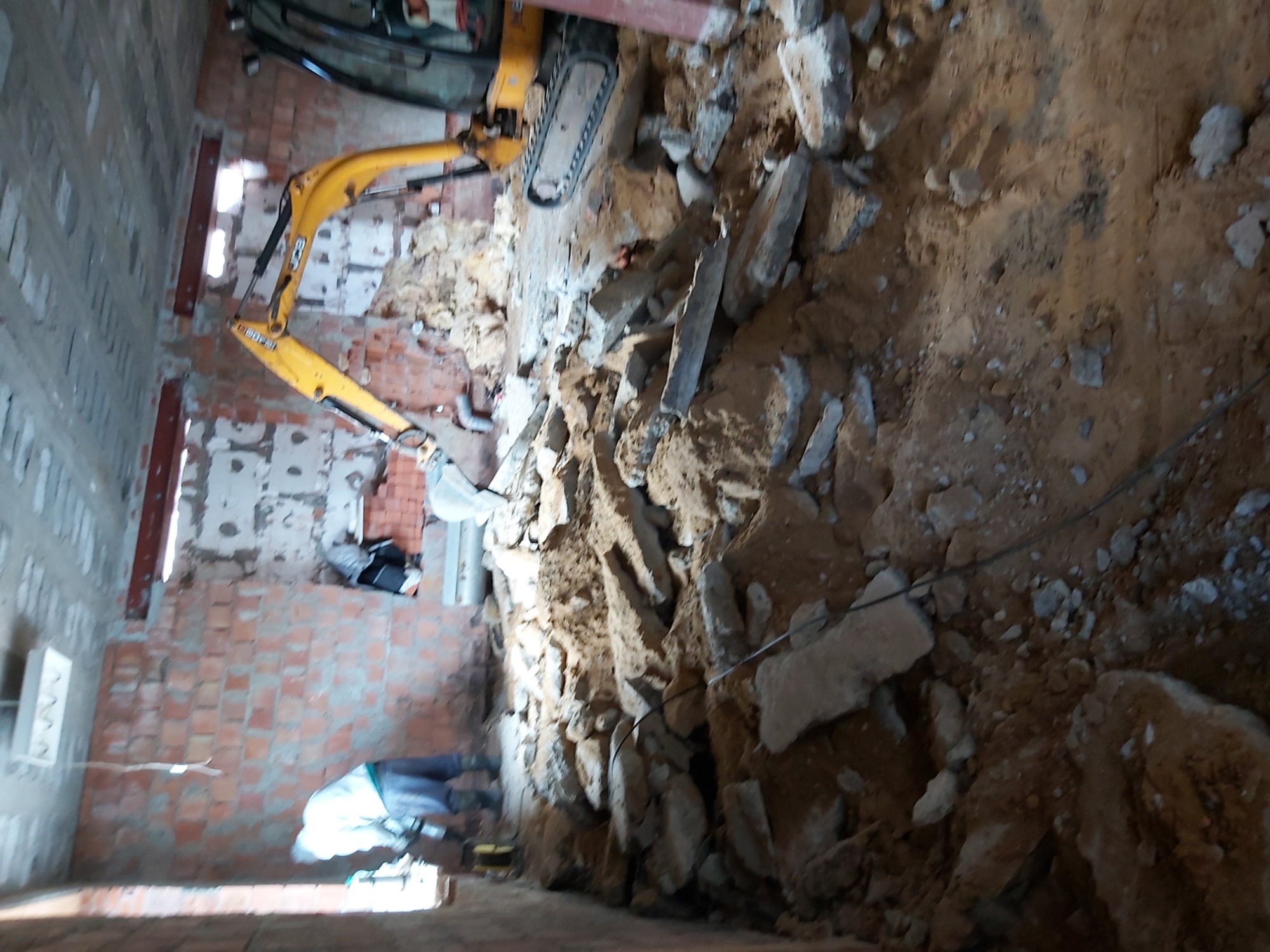 Żółta minikoparka JCB w wykopie wewnątrz budynku z ceglanymi ścianami, widoczne gruz i ziemia.