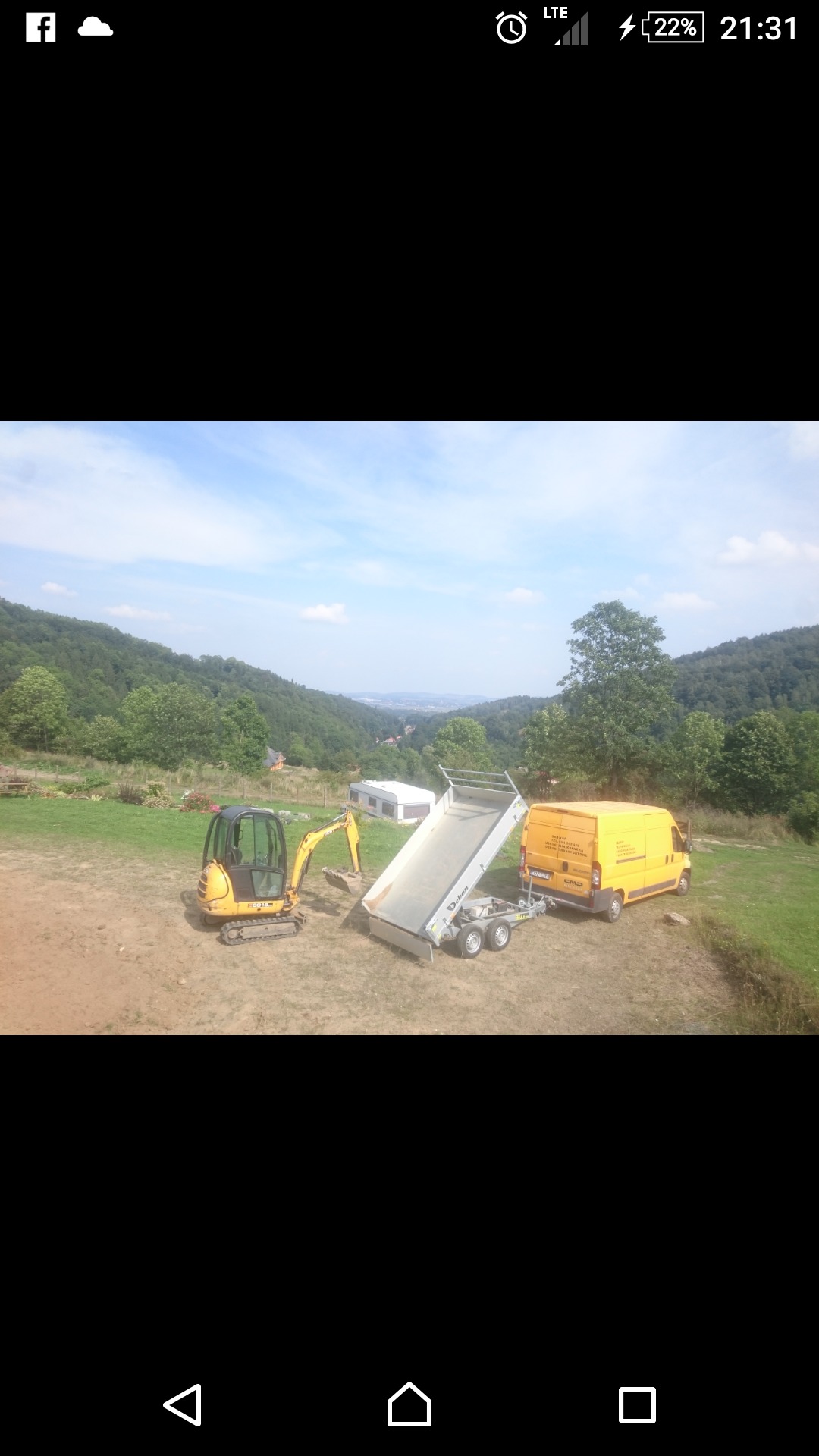 Żółta mini koparka z przyczepą i żółtym vanem na nierównym terenie z widokiem na zalesione wzgórza w tle.