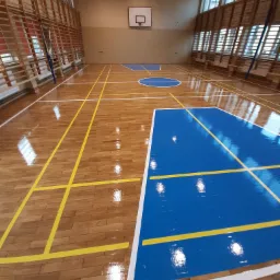 Błyszcząca podłoga w hali sportowej z parkietu, pomalowana na niebiesko w strefach boiska, z żółtymi liniami wyznaczającymi granice, drabinki gimnastyczne przy ścianie.