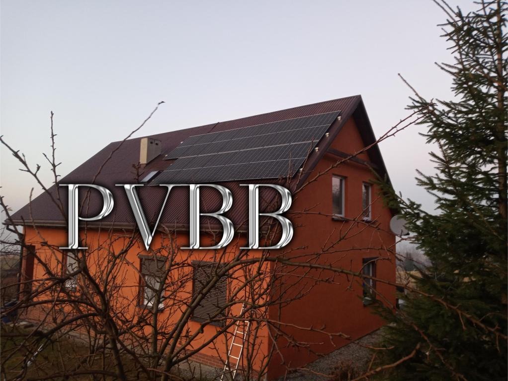 Dom z pomarańczową elewacją i panelami słonecznymi na dachu, widziany zza drzewa z gołymi gałęziami. Na dachu widoczny komin, a z boku domu antena satelitarna.