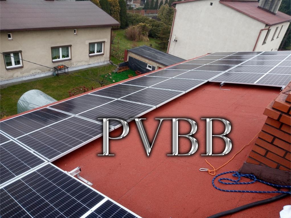 Widok z góry na panele słoneczne zamontowane na dachu pokrytym czerwoną papą, z widocznym kominem z cegły i fragmentem niebieskiej liny zabezpieczającej.