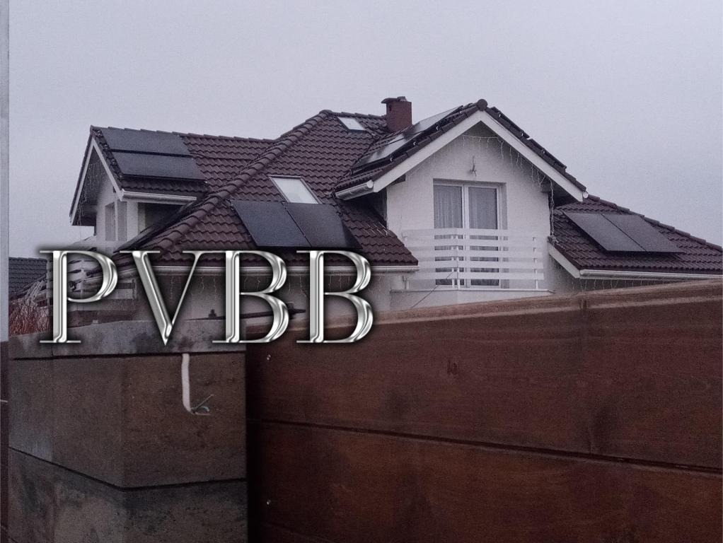 Dom jednorodzinny z brązową dachówką i panelami słonecznymi zamontowanymi na dachu, widoczne zza drewnianego ogrodzenia. Na pierwszym planie napis PVBB.