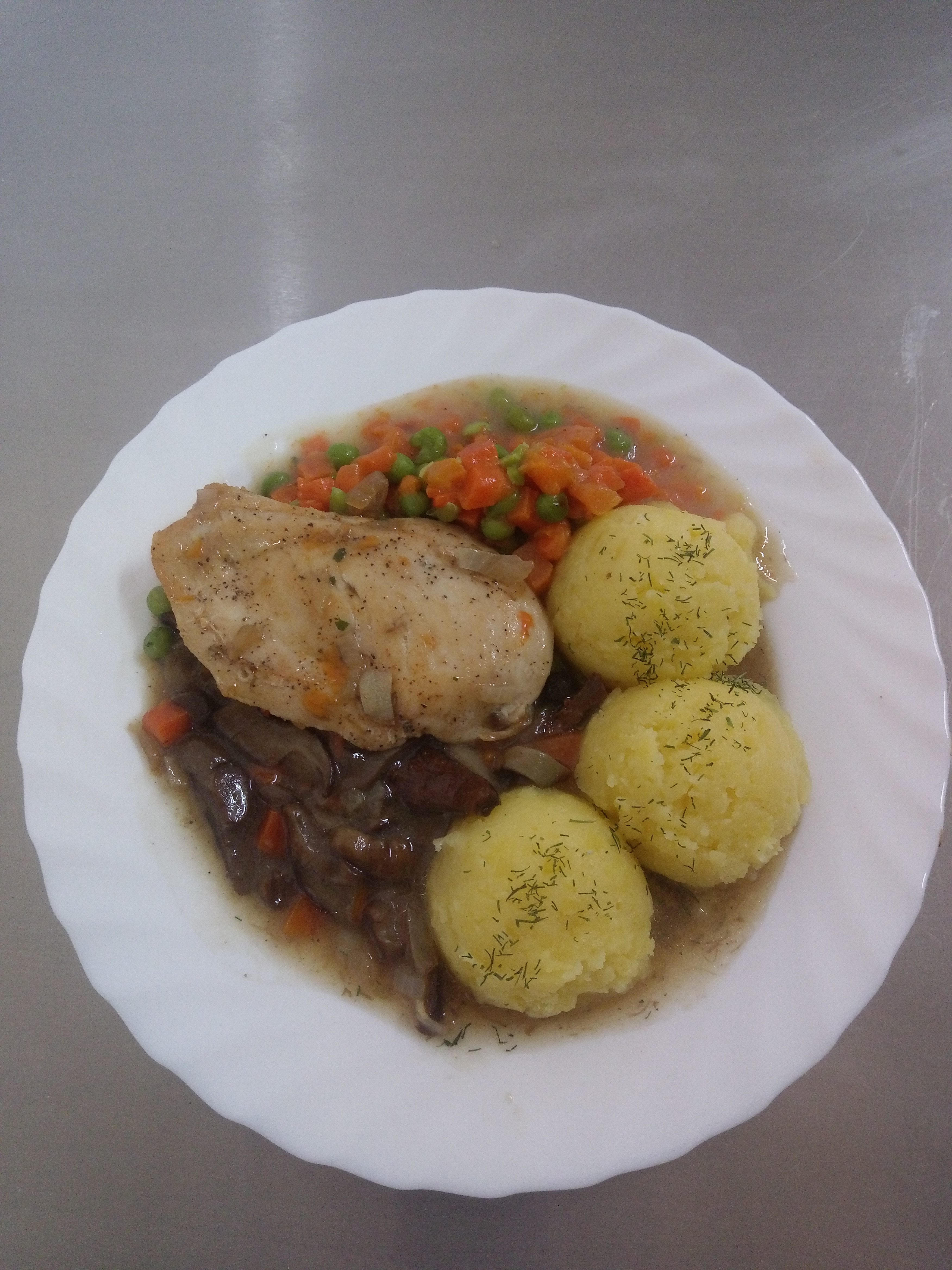 Porcja obiadowa na białym talerzu: pieczony filet z kurczaka z sosem grzybowym, kulki ziemniaczane posypane koperkiem oraz mieszanka gotowanych warzyw (marchewka, groszek) w sosie.