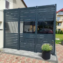 Nowoczesne, poziome panele ogrodzeniowe w kolorze grafitowym, częściowo prześwitujące, oddzielające posesję od sąsiada; w tle widoczny budynek mieszkalny i zaparkowany samochód, a na pierwszym...