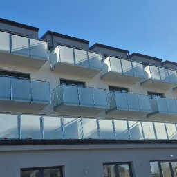 F.H.U.TIG Krzysztof Misiak - Nowoczesny budynek z balkonami zabezpieczonymi szkłem matowym i metalowymi ramami, widziany pod kątem z dołu, na tle błękitnego nieba.