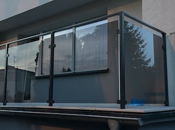 Nowoczesna balustrada szklana z czarnymi profilami na balkonie, widok od dołu, odbicie drzew w szkle.