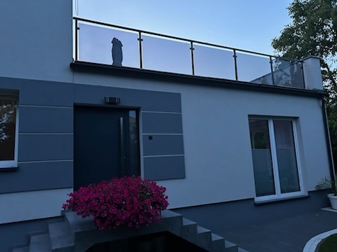 Nowoczesny dom jednorodzinny z balkonem zabezpieczonym szklaną balustradą, na której widać odbicie sylwetki postaci, z wejściem ozdobionym kwiatami.