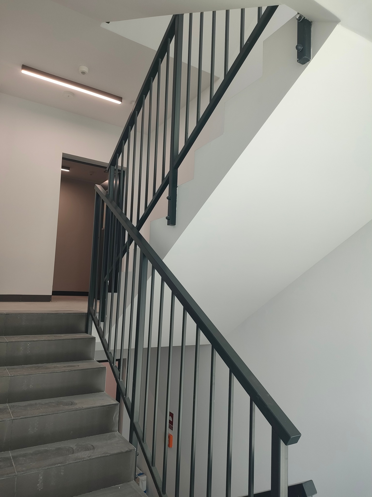 Schody wykończone szarą płytką z ciemną, metalową poręczą w nowoczesnym budynku, widok z dołu na górę biegu schodów.