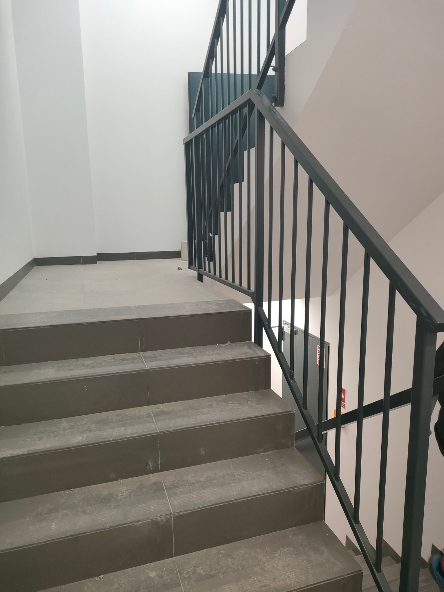 Schody wykończone płytkami z zamontowaną czarną, metalową poręczą. Widok z dołu na górę biegu schodowego.