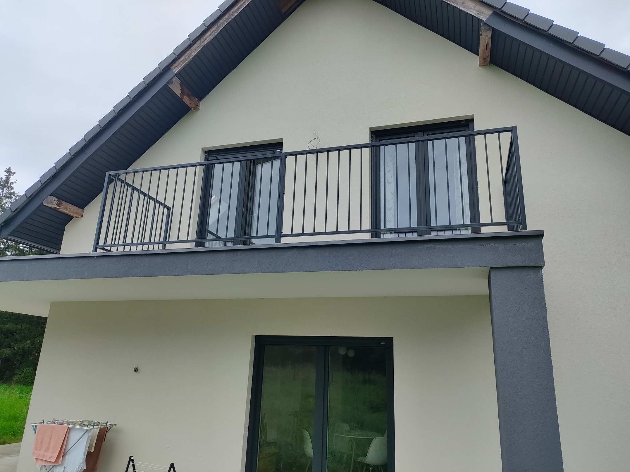 Nowoczesny balkon z czarną, metalową balustradą na tle beżowej elewacji domu z szarym dachem. Na pierwszym planie suszarka na pranie.