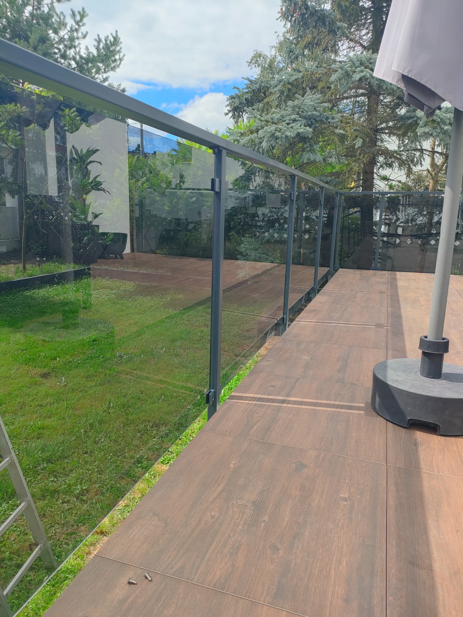 Szklana balustrada tarasowa z czarnymi profilami, widok na ogród, brązowa podłoga z płytek imitujących drewno, częściowo widoczny parasol i drabina.