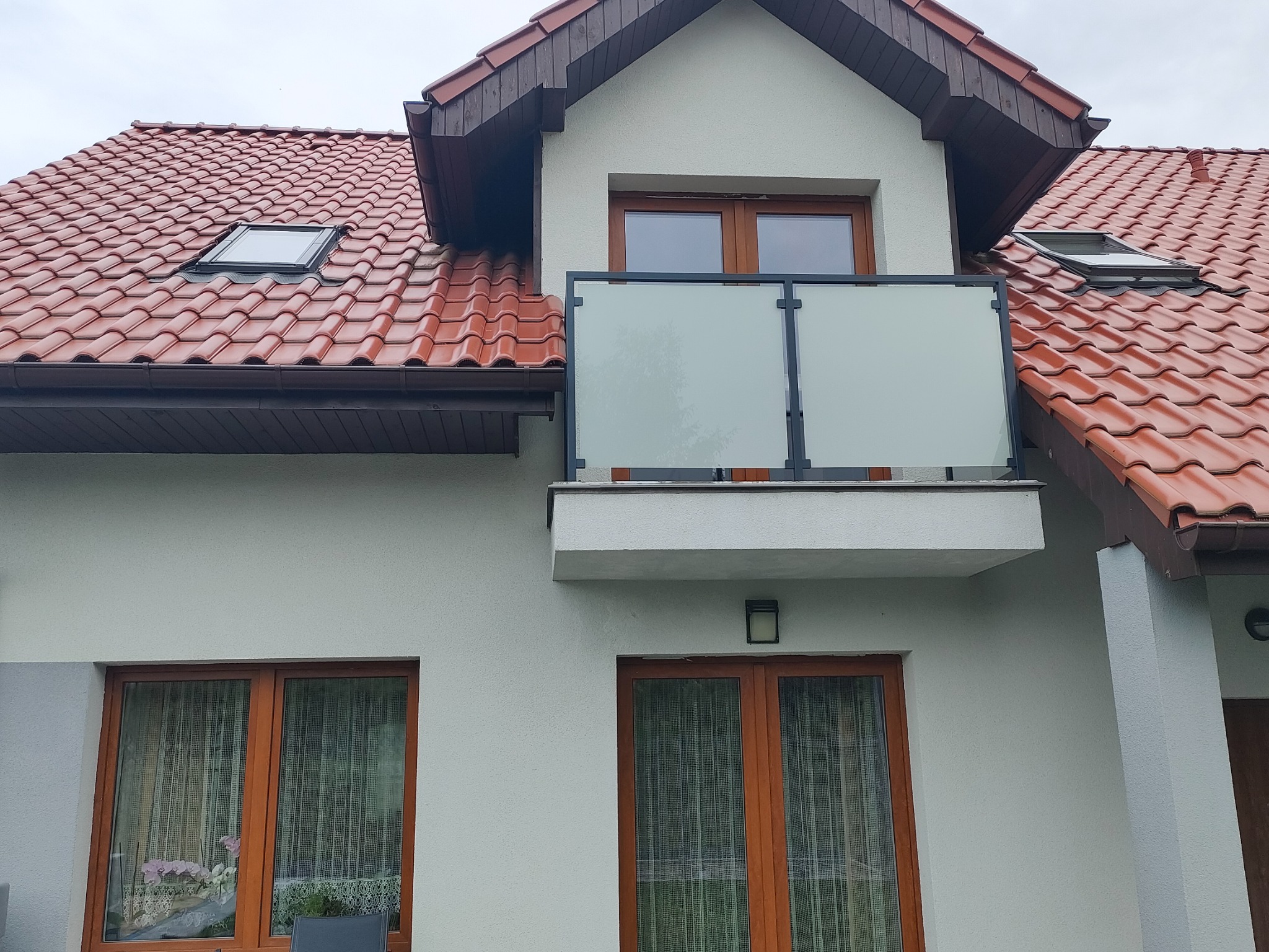 Balkon z matową szybą zamontowaną w czarnej ramie, widok z dołu na budynek z czerwonym dachem i oknami z drewnianą ramą.
