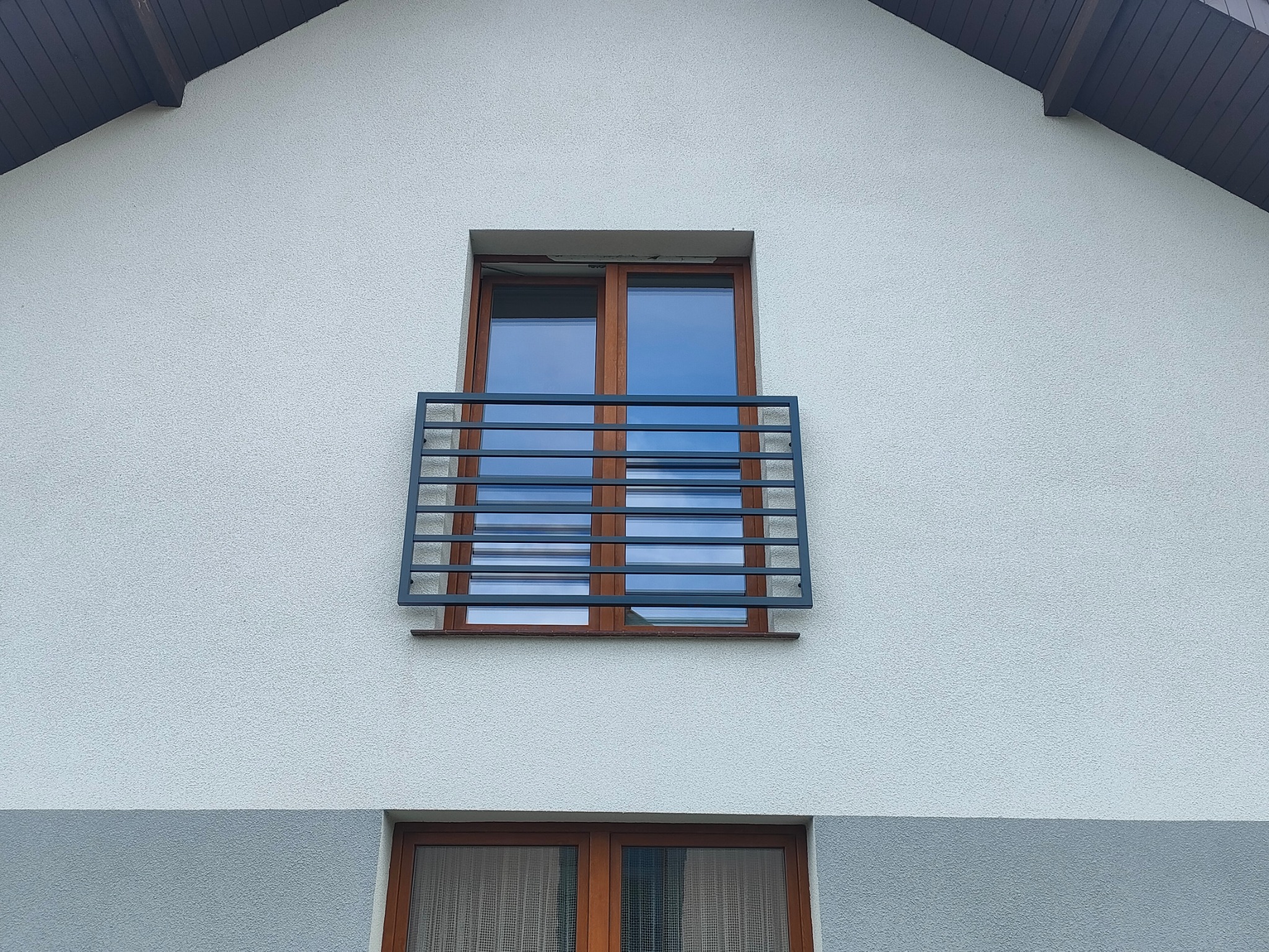 Okienny balkon z szarą, metalową barierką z poziomych profili na tle białej, teksturowanej elewacji domu.