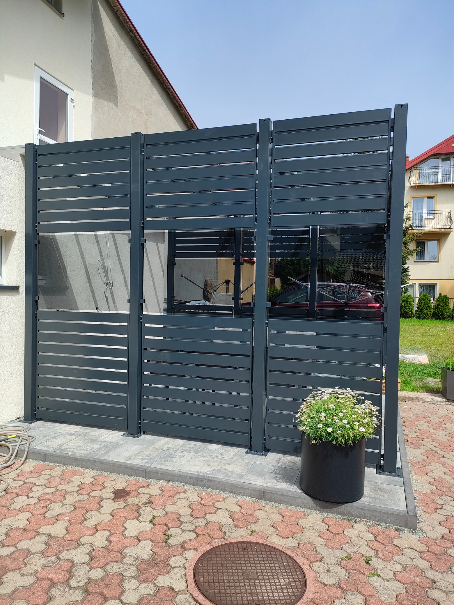 Nowoczesne, poziome panele ogrodzeniowe w kolorze grafitowym, częściowo prześwitujące, oddzielające posesję od sąsiada; w tle widoczny budynek mieszkalny i zaparkowany samochód, a na pierwszym...
