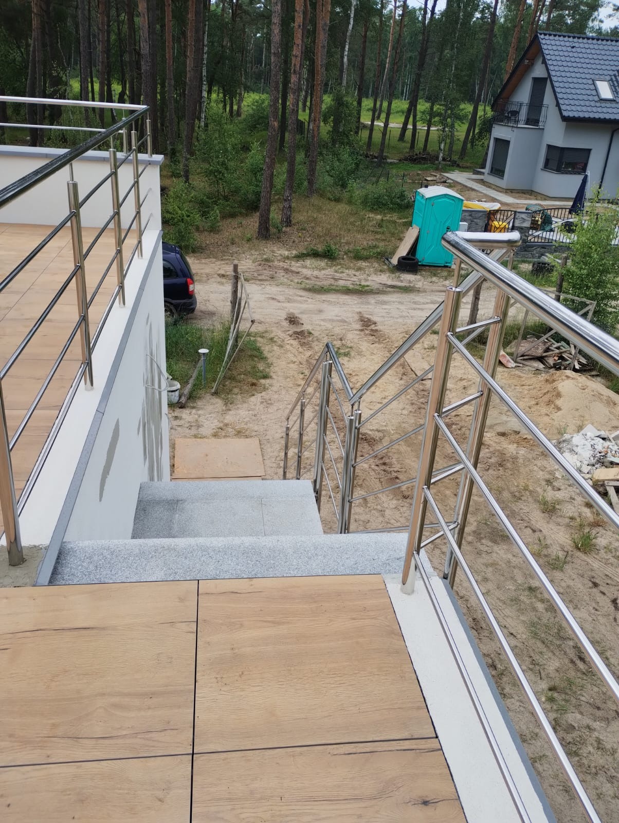Zewnętrzne schody z szarego granitu prowadzące z tarasu wyłożonego drewnopodobnymi płytkami, zabezpieczone metalową poręczą z nierdzewnej stali, w tle dom jednorodzinny i las.