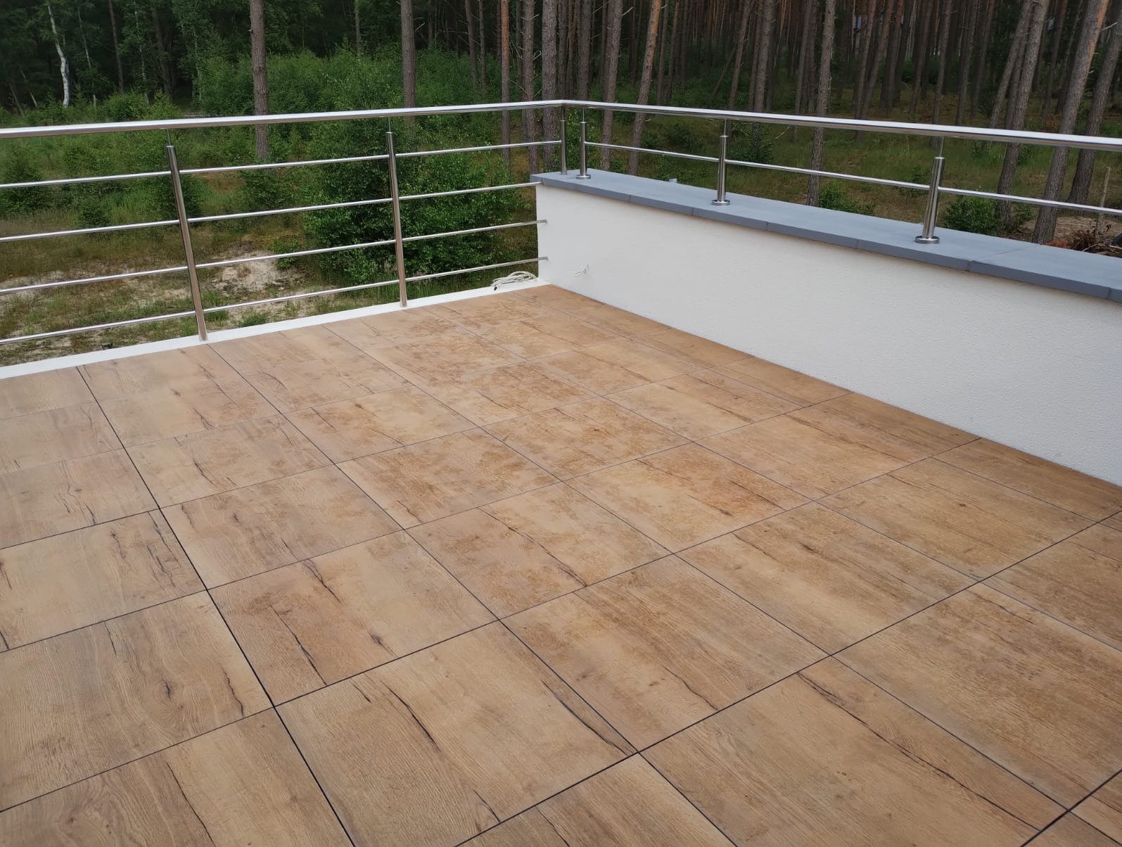 Balkon z brązowymi płytkami imitującymi drewno i stalową balustradą z widokiem na las.