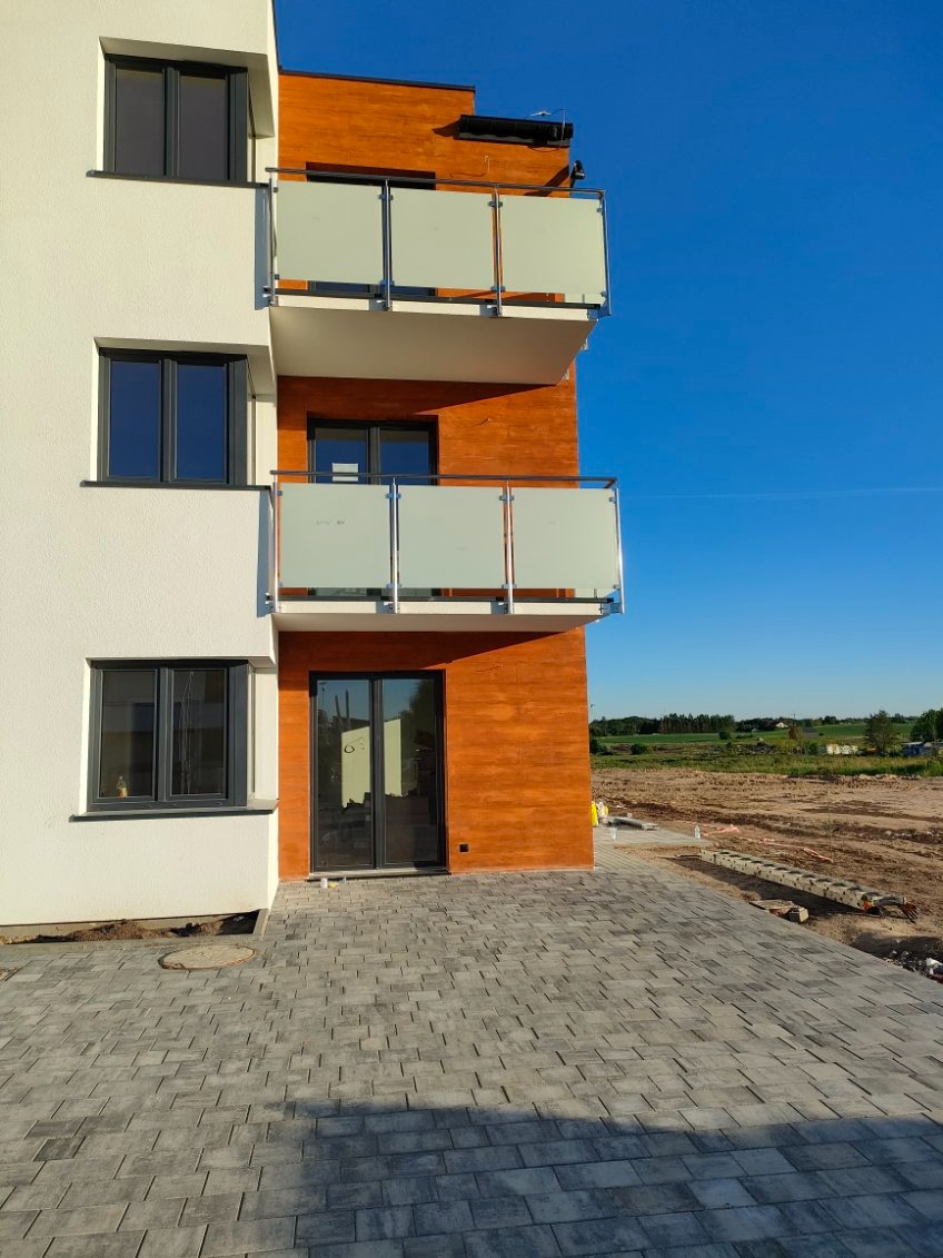 Nowoczesny budynek z balkonami zabezpieczonymi szkłem mlecznym osadzonym w metalowych ramach, widok z poziomu brukowanego podjazdu, w tle plac budowy.