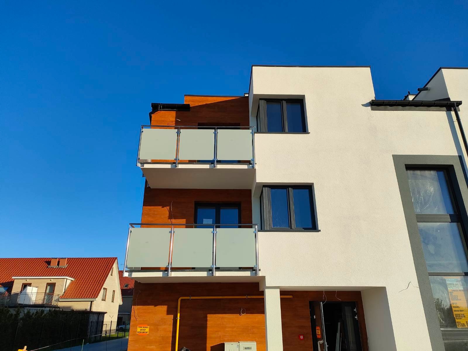 Nowoczesny budynek z balkonami zabezpieczonymi szkłem hartowanym i stalowymi ramami, widoczne instalacje gazowe na elewacji.
