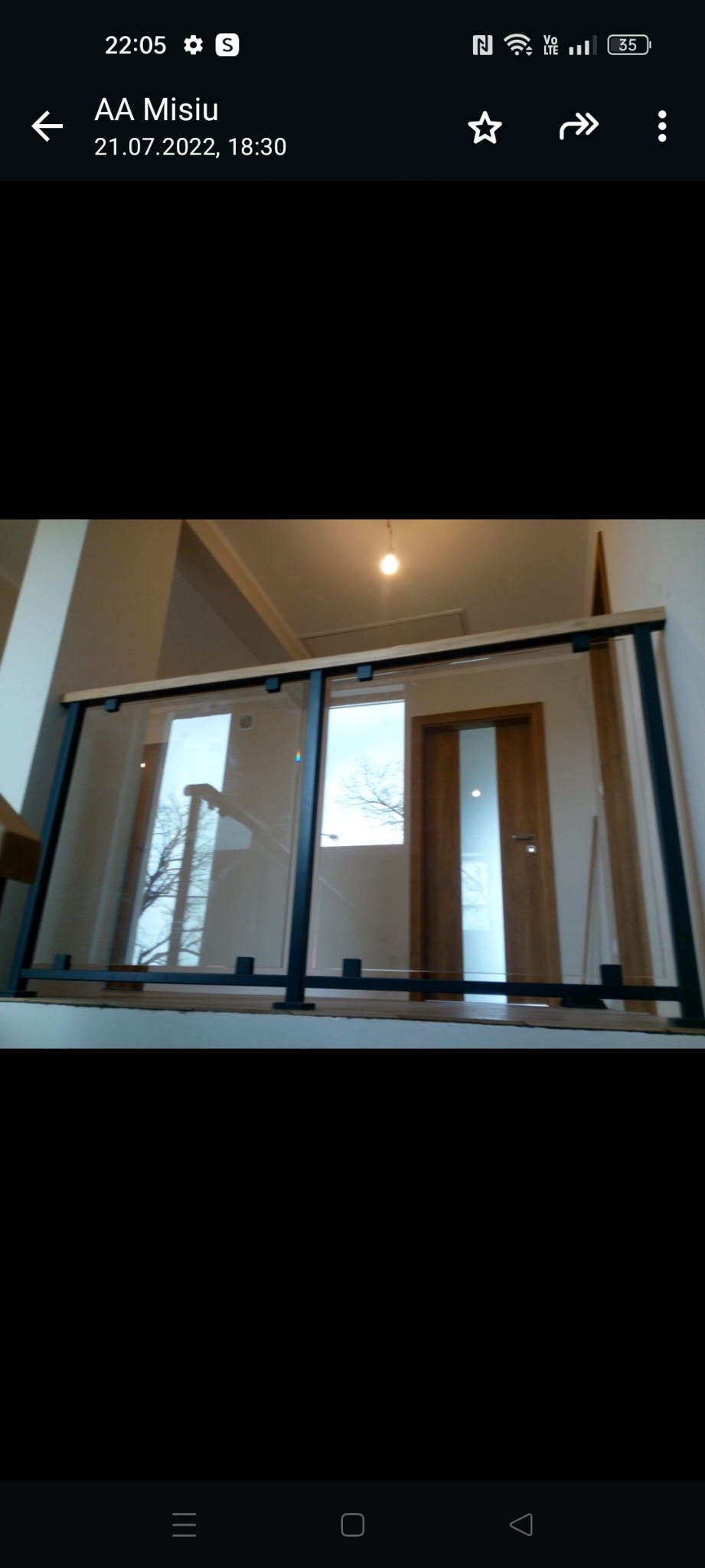 Szklana balustrada z czarnymi elementami mocującymi i drewnianą górną poręczą, widok z dołu na fragment antresoli z drzwiami i oknem z widokiem na drzewa.