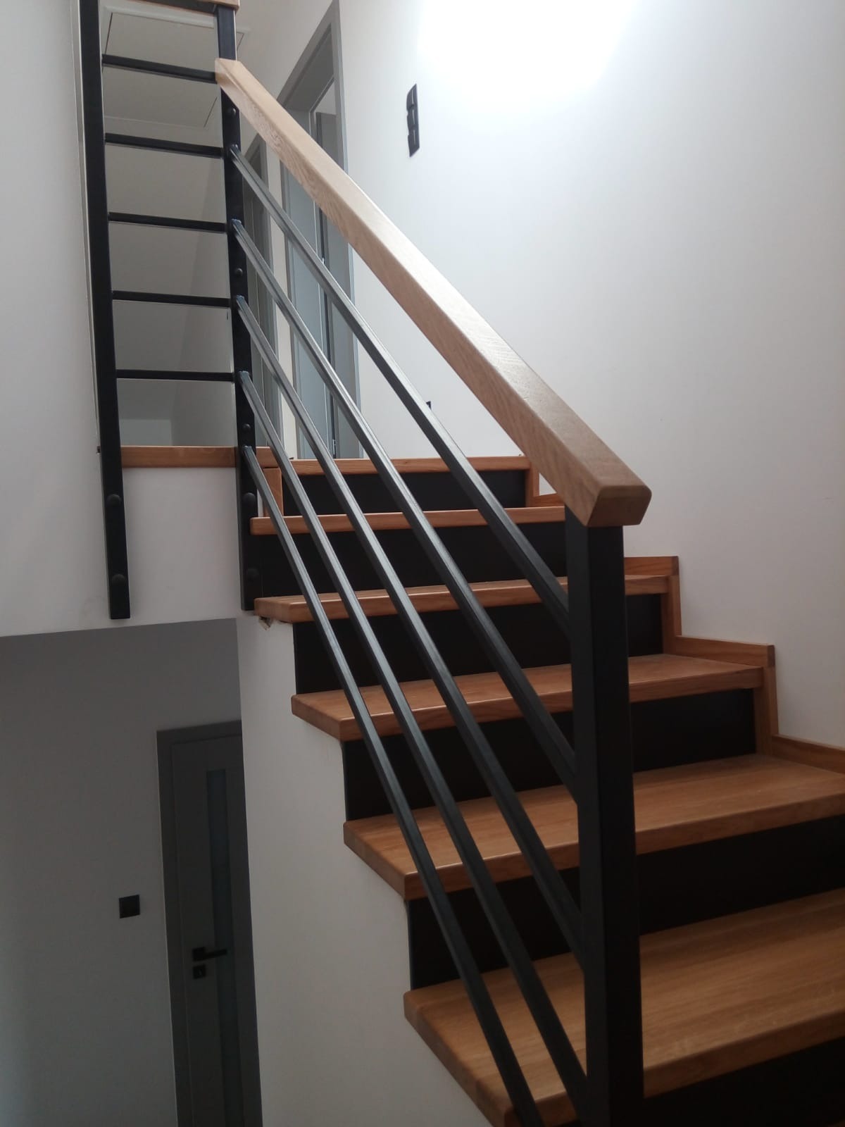 Schody z drewnianymi stopniami i czarną, metalową poręczą z drewnianym uchwytem, widok z dołu na jasne wnętrze.