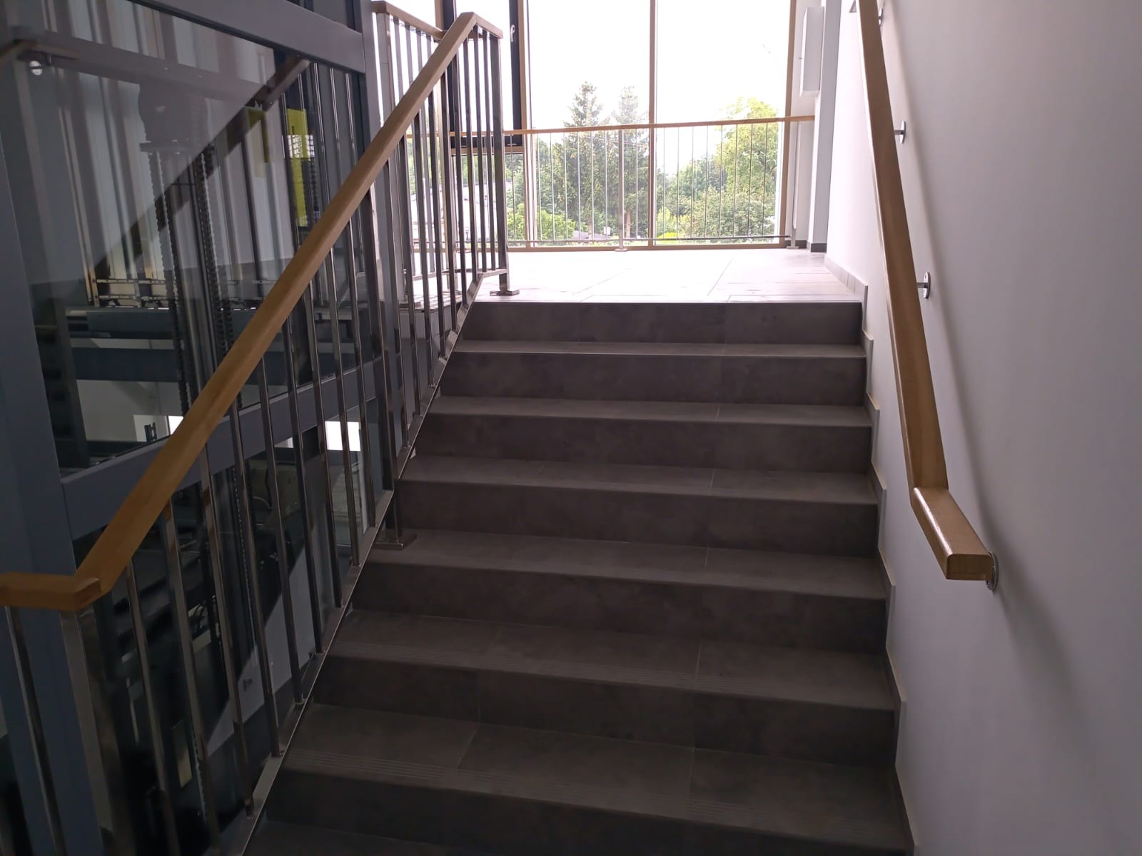 Schody z szarych płytek z poręczą z jasnego drewna po prawej stronie i balustradą ze stali nierdzewnej po lewej, widok z dołu na górę, z oknem na końcu schodów.