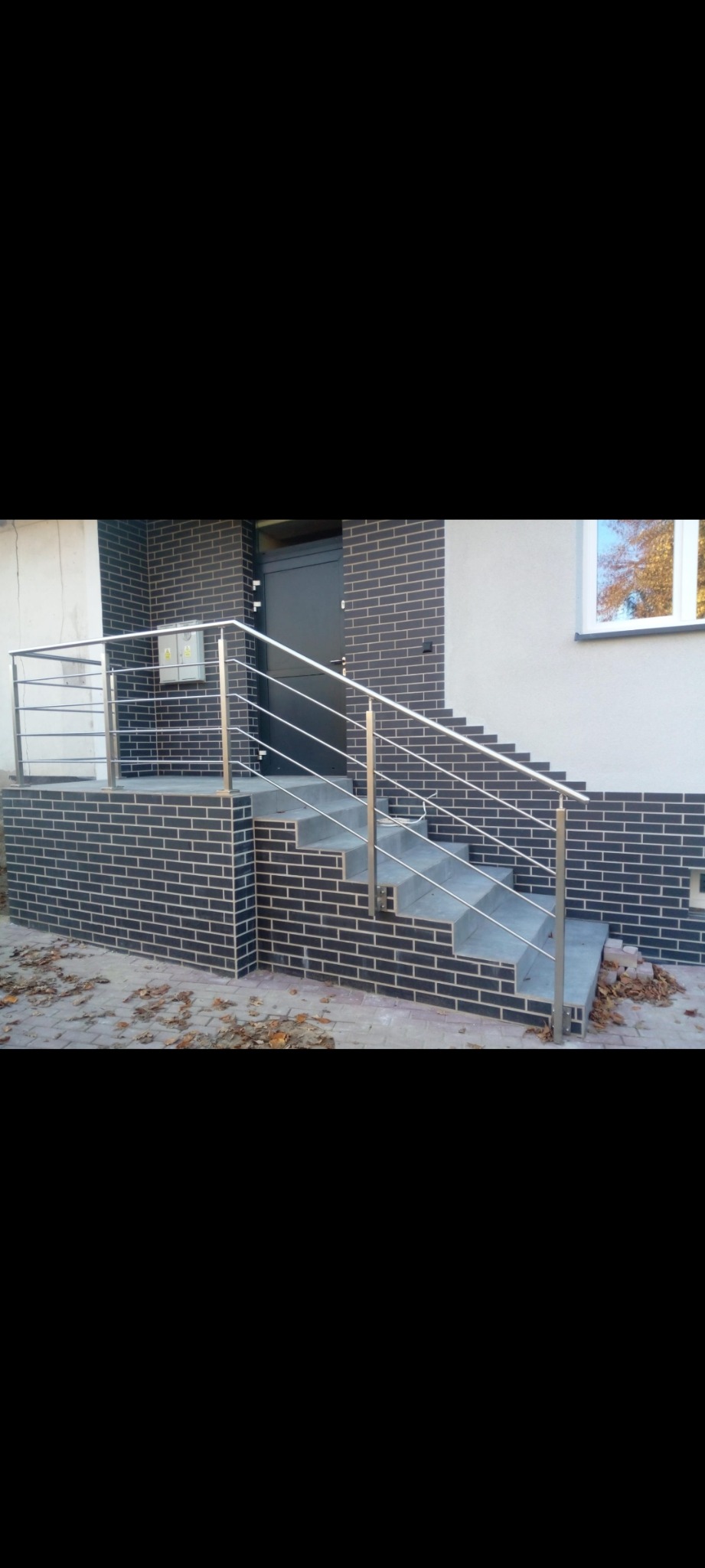 Zewnętrzne schody wejściowe do budynku z metalową poręczą z prostych rurek, wykończone ciemną cegłą klinkierową i szarymi stopniami.