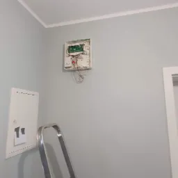 Otwarta obudowa centrali alarmowej z wystającymi przewodami na szarej ścianie, widoczna drabina aluminiowa i skrzynka elektryczna