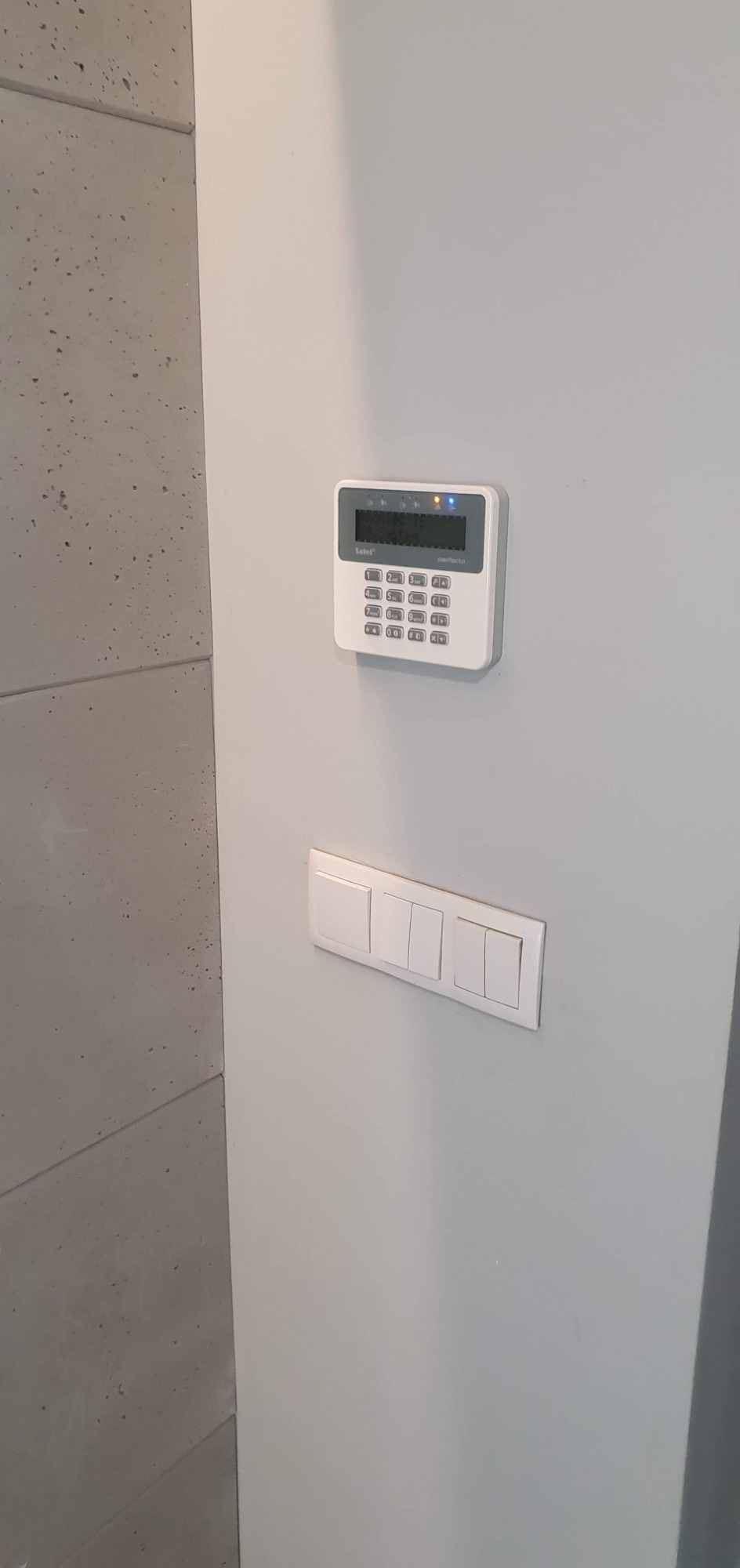 Centrala alarmowa Satel Perfecto z podświetlonym wyświetlaczem i klawiaturą numeryczną, zamontowana na szarej ścianie obok włączników światła.