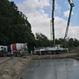 Wylewanie betonu na przygotowany zbrojony fundament za pomocą pompy do betonu CIFA, widoczna betoniarka z czerwono-biało-niebieskim bębnem oraz pracownicy w żółtej kamizelce i bez koszulki.