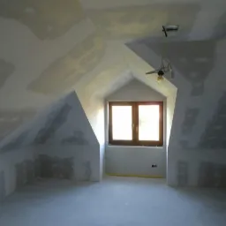 Poddasze z oknem w szczycie, ściany i sufit w trakcie szpachlowania, widoczne łączenia płyt gipsowo-kartonowych, surowa żarówka zwisająca z sufitu.