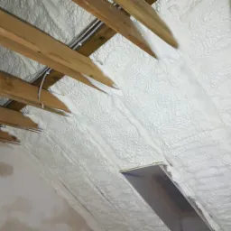 Poddasze z drewnianą konstrukcją krokwiową pokryte grubą warstwą białej piany izolacyjnej, widoczny fragment okna dachowego.