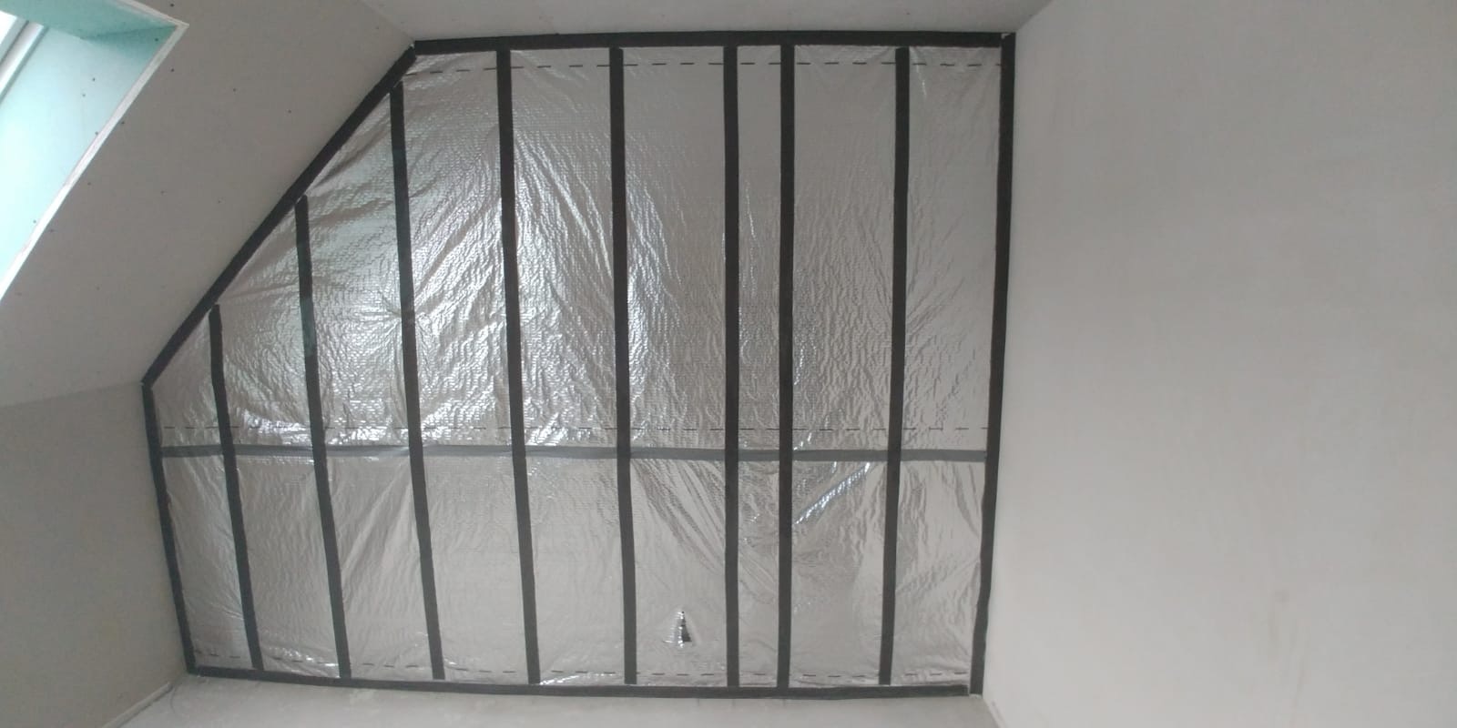 Srebrna folia izolacyjna zamontowana na czarnych profilach w skośnym poddaszu, widoczne elementy okna dachowego, etap prac przed montażem płyt kartonowo-gipsowych.
