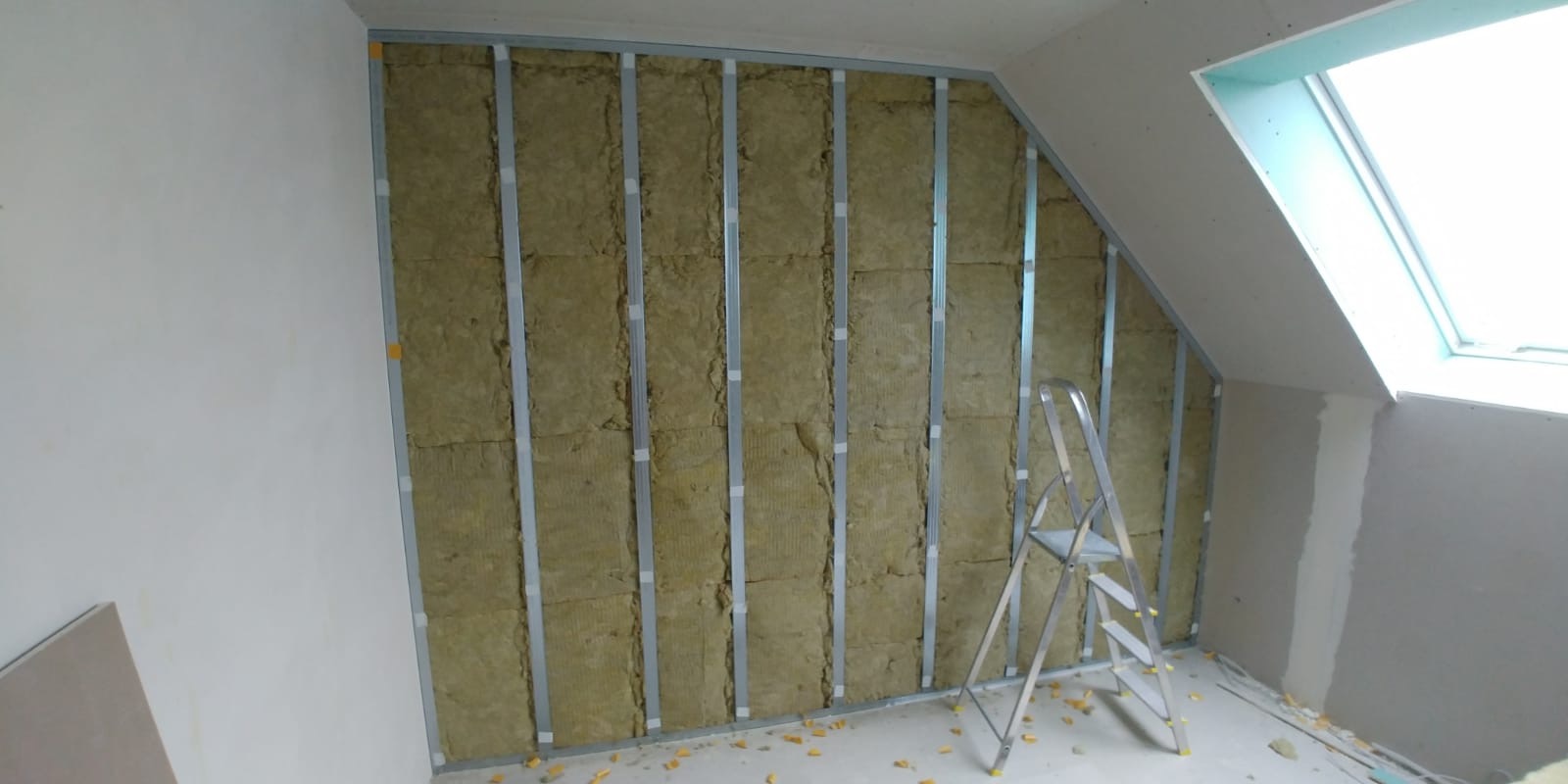 Skośne poddasze w trakcie izolacji: widoczne metalowe profile, wełna mineralna między nimi, drabina aluminiowa i okno dachowe.