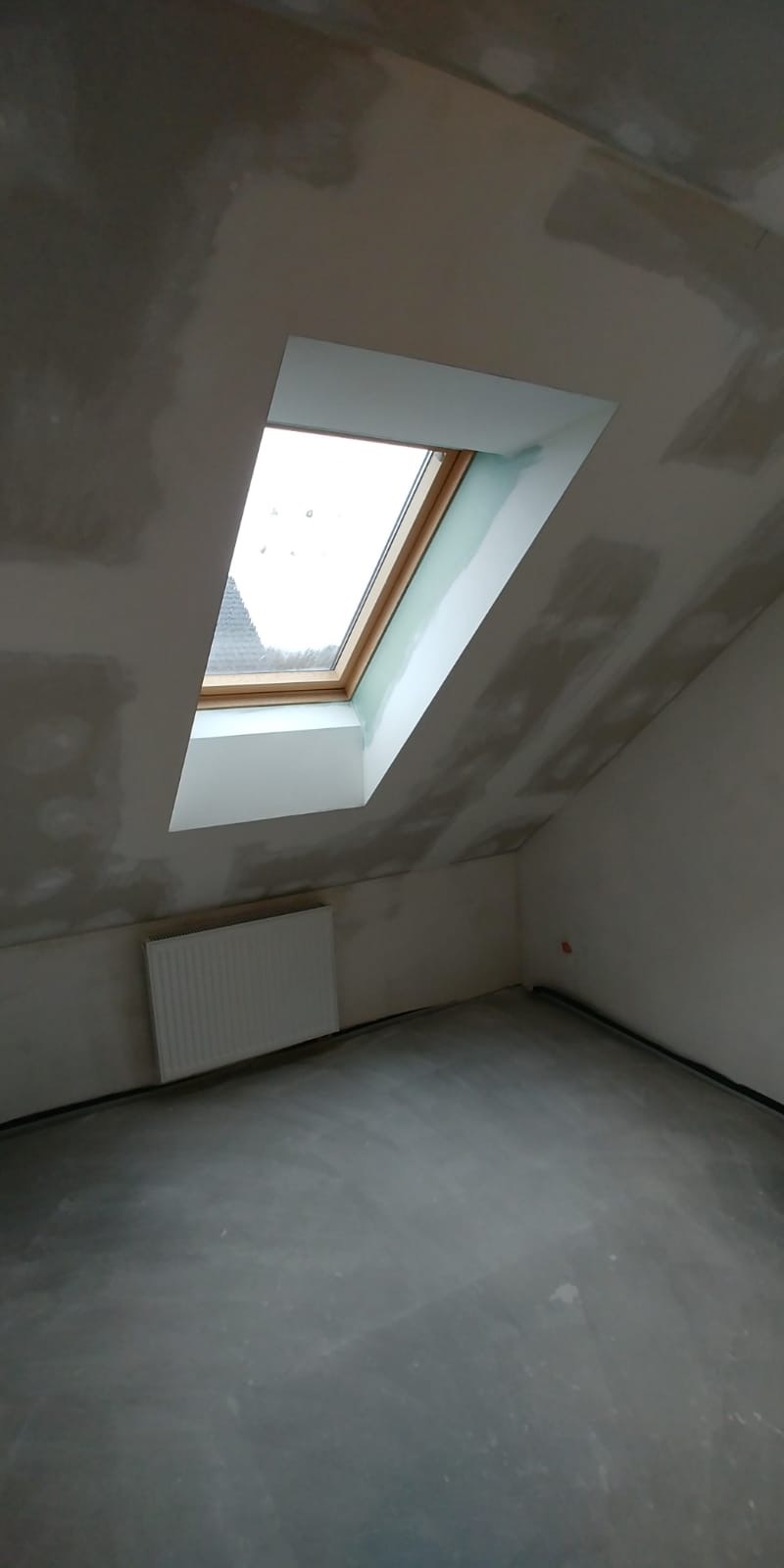 Poddasze w trakcie wykańczania: widoczne okno dachowe z drewnianą ramą, grzejnik, ściany z widocznymi śladami szpachlowania i surowa betonowa podłoga.