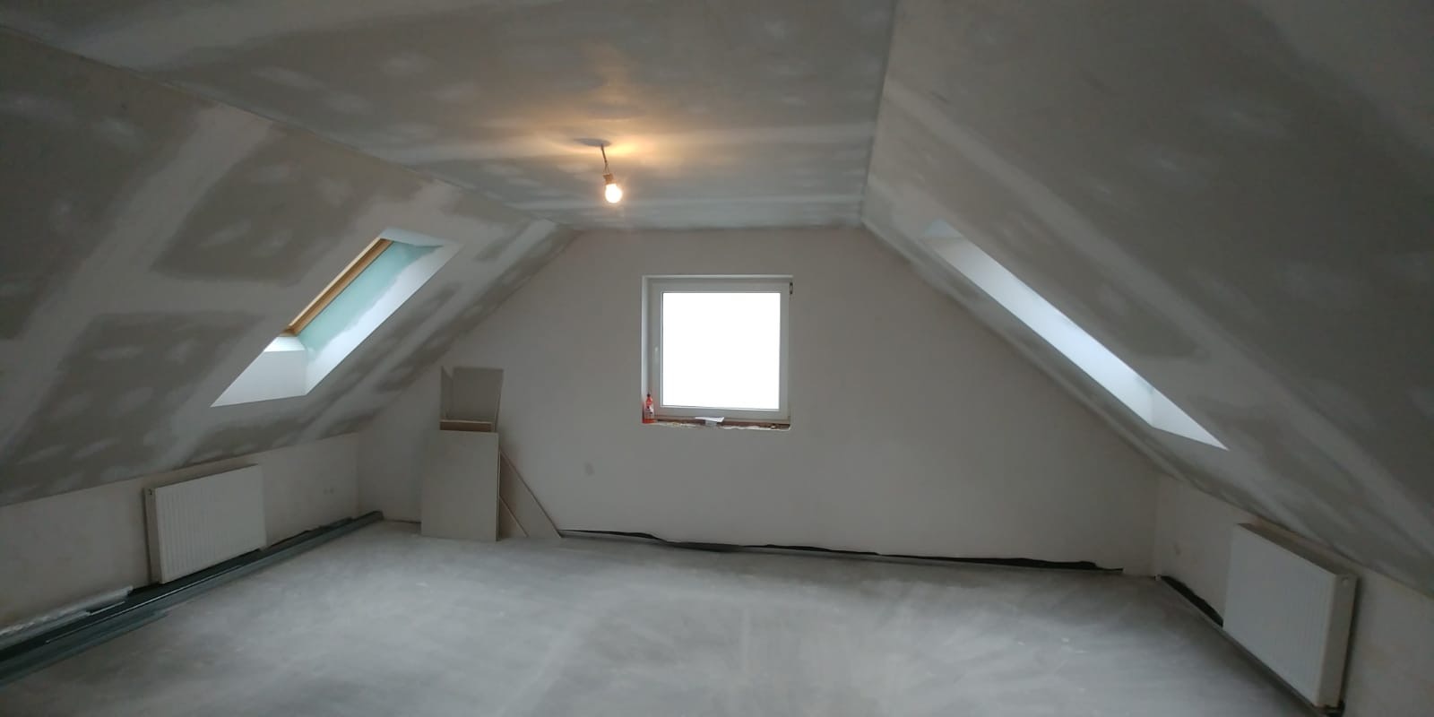 Poddasze w trakcie wykańczania: widoczne okna dachowe, grzejniki, częściowo położone płyty gipsowo-kartonowe na skosach i suficie, oraz surowa podłoga.
