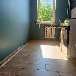 Ukończona podłoga z płytek imitujących drewno w niebieskiej kuchni, listwy przypodłogowe częściowo zamontowane, widoczny grzejnik i kuchenka, wpadające światło słoneczne.