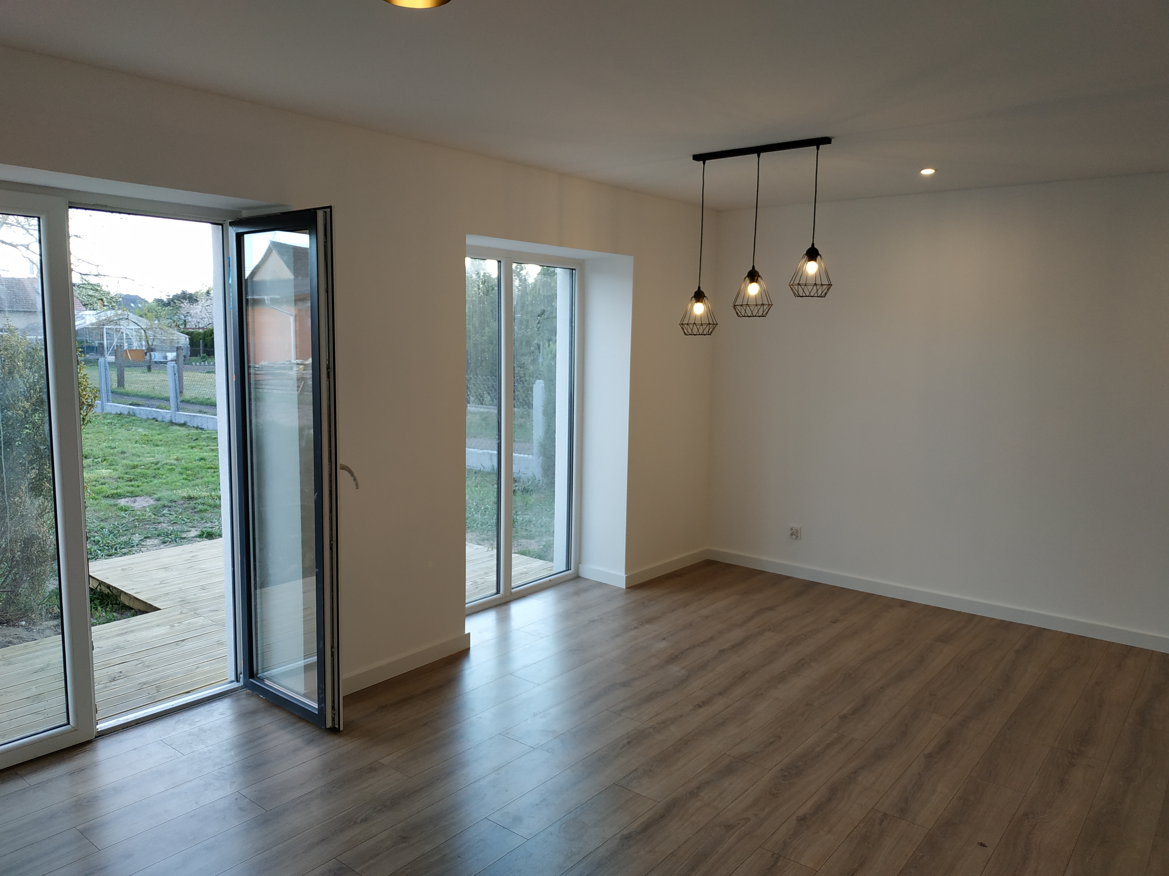Jasne, przestronne wnętrze z drewnianą podłogą, białymi ścianami, trzema wiszącymi lampami w stylu industrialnym i otwartymi drzwiami balkonowymi z widokiem na ogród.