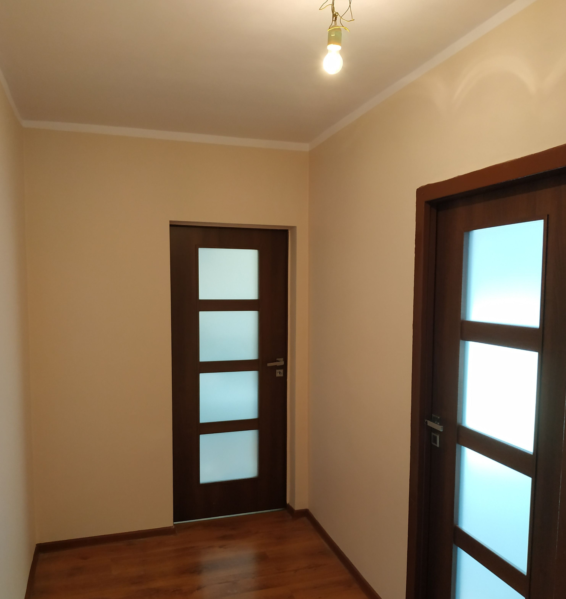 Wnętrze po remoncie w Opolu: Dwie nowoczesne, ciemnobrązowe drzwi z matowymi szybami, świeżo pomalowane ściany w odcieniach beżu, parkiet na podłodze, widoczna żarówka na suficie.