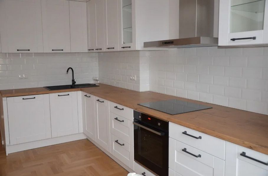 Nowoczesna kuchnia z białymi szafkami, drewnianym blatem i czarnymi uchwytami, wykończona białą cegiełką na ścianie i parkietem na podłodze.
