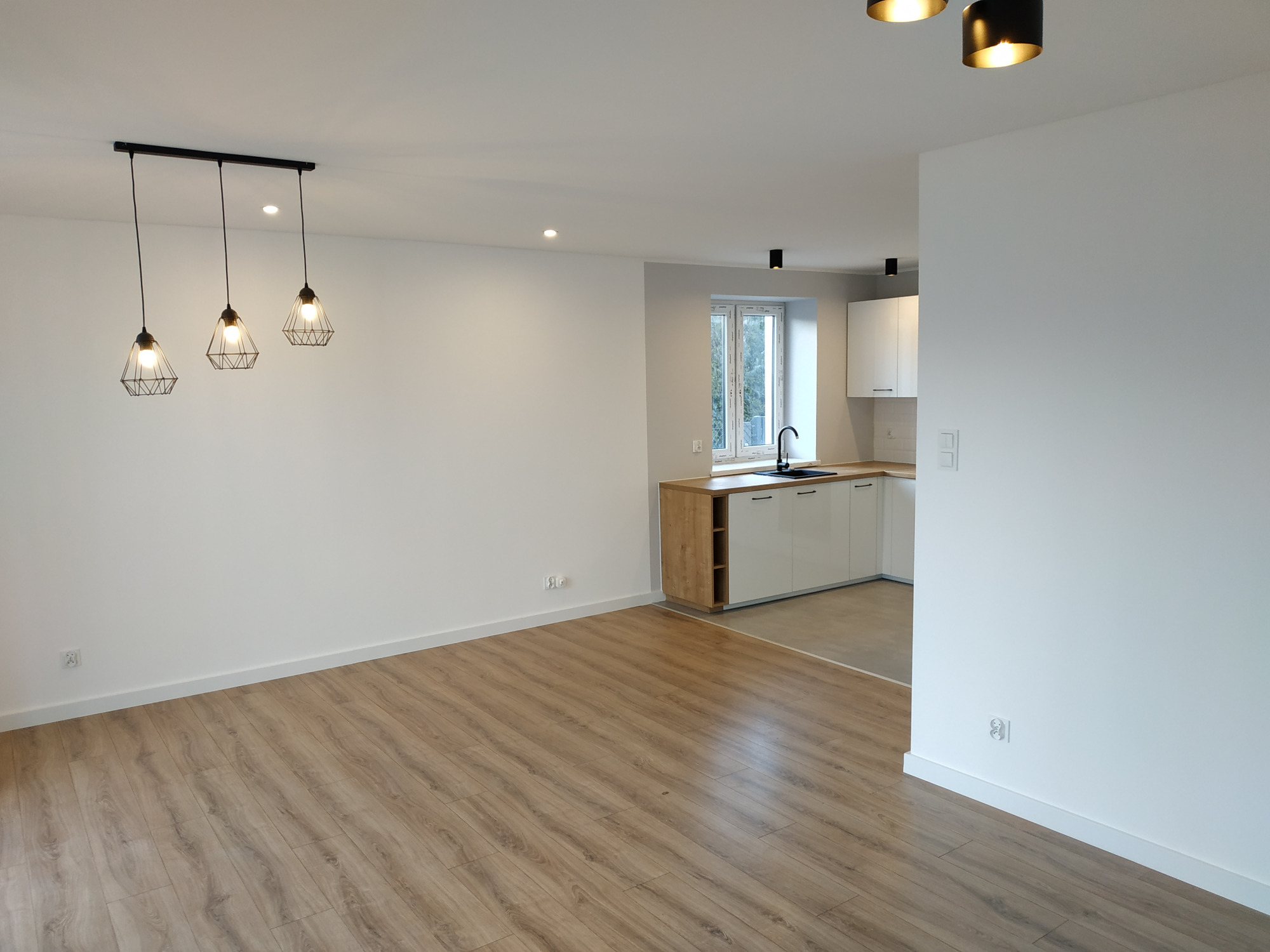 Jasne, przestronne wnętrze po remoncie: panele podłogowe w odcieniach drewna, białe ściany, nowoczesne oświetlenie z trzema wiszącymi lampami o geometrycznych kloszach oraz widoczna kuchnia...
