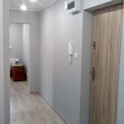 Korytarz po remoncie z szarymi ścianami, panelami podłogowymi imitującymi drewno i drewnianymi drzwiami. Na ścianie widoczny biały domofon i skrzynka elektryczna.