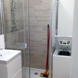 Wasrem - Nowoczesna łazienka z kabiną prysznicową z przesuwnymi drzwiami, białą umywalką z szafką, pralką z otwartym bębnem oraz akcesoriami do sprzątania w rogu kabiny.