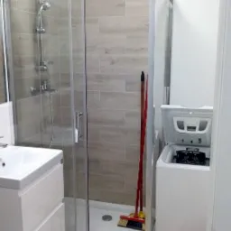 Nowoczesna łazienka z kabiną prysznicową z przesuwnymi drzwiami, białą umywalką z szafką, pralką z otwartym bębnem oraz akcesoriami do sprzątania w rogu kabiny.