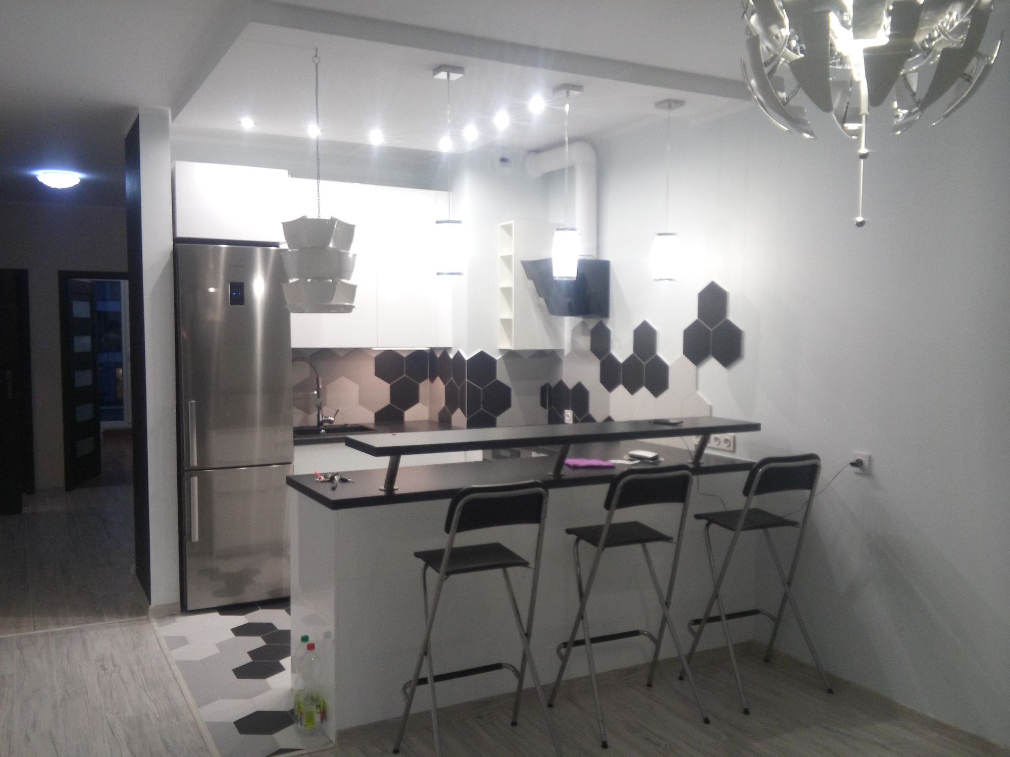 Nowoczesna kuchnia z białymi szafkami, stalową lodówką, czarnym blatem barowym i heksagonalnymi płytkami na ścianie oraz podłodze.