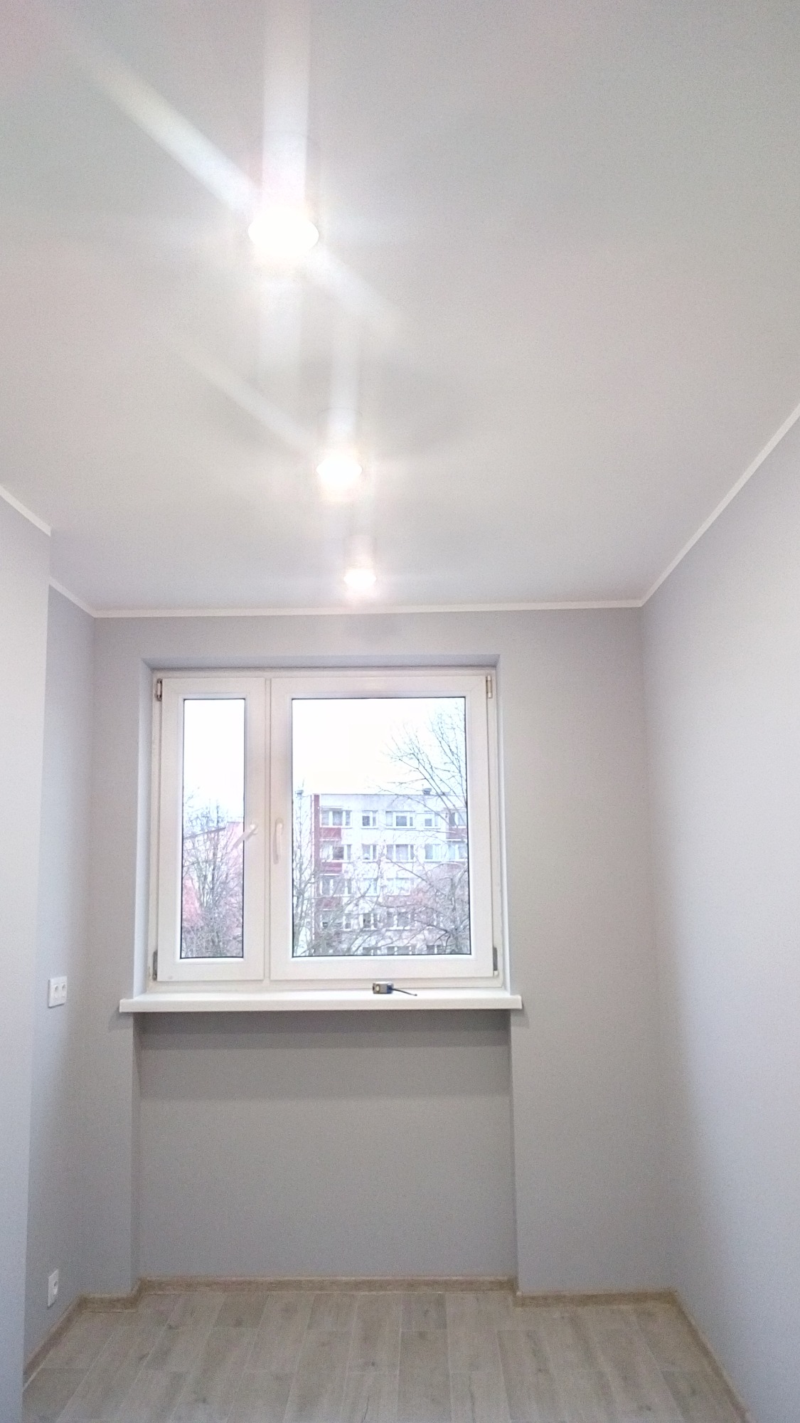 Pomalowane na szaro wnętrze pokoju z oknem, jasnymi panelami podłogowymi i trzema punktowymi lampami sufitowymi.